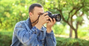 A man wearing a denim jacket is outdoors, holding a camera up to his face and taking a photograph, with green trees and bushes blurred in the background.